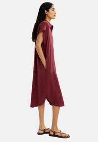 Burgundowa, oversize sukienka z miękkiego materiału, z kołnierzykiem, krótkimi rękawami, bocznymi kieszeniami i zaokrąglonym dołem. Noszona z brązowymi sandałami.