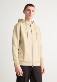 Felpa beige con zip e cappuccio regolabile, tasche anteriori e polsini a costine. Tessuto liscio con vestibilità comoda, abbinata a pantaloni scuri.