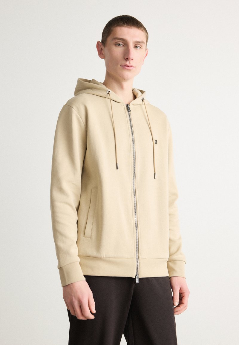 Felpa beige con zip e cappuccio regolabile, tasche anteriori e polsini a costine. Tessuto liscio con vestibilità comoda, abbinata a pantaloni scuri.