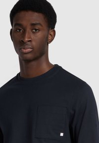 Marineblaues Langarm-T-Shirt aus Baumwolle mit Brusttasche und Etikett, minimalistisches Design und runder Ausschnitt.