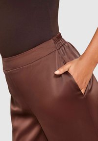 Hand rustend in de zak van glanzende, bruine satijnen broeken met een elastische tailleband, gecombineerd met een aansluitend donkerbruine top tegen een effen achtergrond.