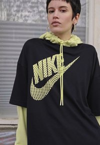 Musta ülisuure Nike t-särk kollase võrkmaterjali logoga ja kummipaelaga kapuuts. Omab lühikesi varrukaid ja lõdva lõiget, kihiliselt heledat t-särki.