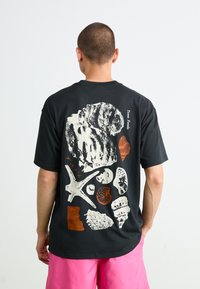 Mand iført en sort t-shirt med hvide og orange illustrationer af fossiler og skaller samt teksten "Texas Fossils" på ryggen, paret med klare pink shorts.