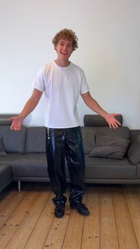 Μαύρο γυαλιστερό βινύλιο παντελόνι και ένα απλό λευκό κοντομάνικο μπλουζάκι, στέκεται πάνω σε ξύλινο πάτωμα με έναν γκρίζο καναπέ στο παρασκήνιο.