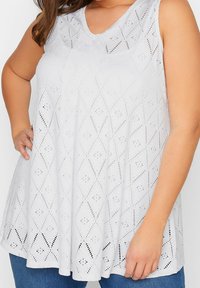 Ärmlös vit blus i ögonlåt med diamants- och prickmönster, bärs av en person med handen på höften och blå jeans.