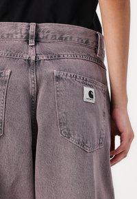 Violet džinsai su aukštu juosmeniu, su dviem užpakalinėmis kišenėmis ir Carhartt etikete, siuvinėta dešinėje kišenėje. Tekstūruota medžiaga.