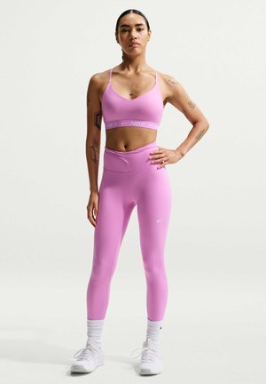 Αθλητικό άτομο στέκεται με αυτοπεποίθηση φορώντας ροζ αθλητικό σουτιέν Nike και λεγινέτες με λευκά αθλητικά παπούτσια και κάλτσες σε απλό φόντο.