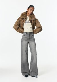 Jachetă bomber maro cu guler din blană, tricou crem cu logo, blugi gri cu talie înaltă și picior larg, și pantofi cu toc ascuțit negri.