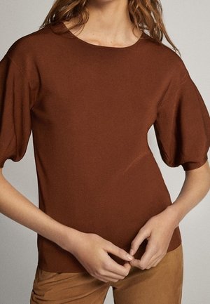 Kobieta ubrana w brązową bluzkę z krótkim rękawem z bufkami oraz dopasowane brązowe spodnie, stojąca ze złożonymi dłońmi.