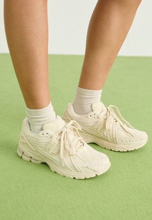 Benen met witte sokken en gebroken witte New Balance chunky sneakers staan op een lichtgroene vloer.