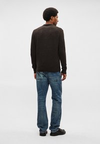 Σκούρο καφέ πλεκτό πουλόβερ με στρογγυλή λαιμόκοψη, συνδυασμένο με ανοιχτό μπλε τζιν. Η υφή φαίνεται απαλή και η εφαρμογή είναι χαλαρή.