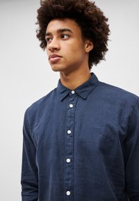 Camisa de manga larga azul marino hecha de tejido ligero, con un cuello clásico, cierre delantero con botones y un sutil logo bordado en el pecho.