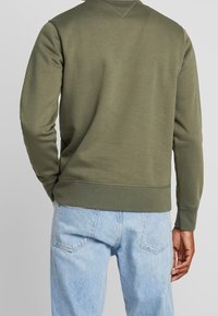 Sudadera verde oliva con mangas largas, puños y dobladillo acanalados. Textura suave y diseño sin cremallera. Combinada con jeans de color azul claro.