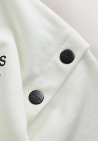 Tissu blanc à texture lisse avec un bouton noir gravé de "ALLSAINTS", positionné à la couture de l'épaule d'un vêtement.