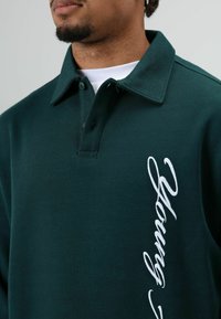 Donkergroen poloshirt van katoen. Heeft een kraag, knoopsluiting en witte geborduurde tekst aan de linkerkant.