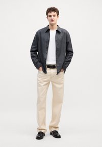 Camicia button-up grigio scuro, t-shirt a girocollo bianca sotto, pantaloni color crema e scarpe nere; il modello sta in piedi con le mani in tasca.