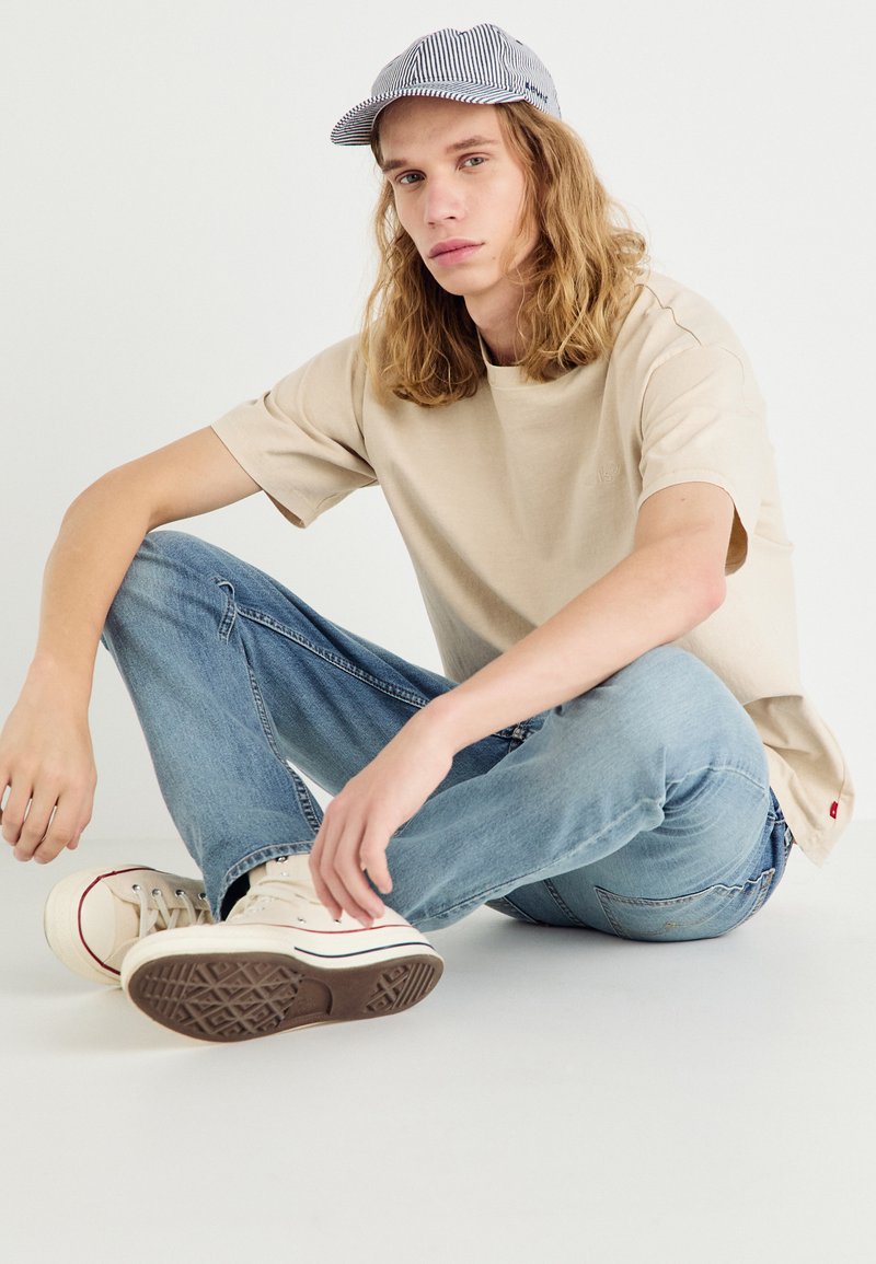 T-shirt en coton beige, jean en denim bleu clair et baskets crème avec accents rouges. Casquette à rayures grises et blanches. Modèle assis sur un fond blanc.