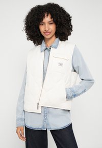 Žena s kovrčavom kosom nosi bijelu prešanu prsluk preko svijetle traper košulje i tamnih traperica, stojeći ispred jednostavne pozadine.