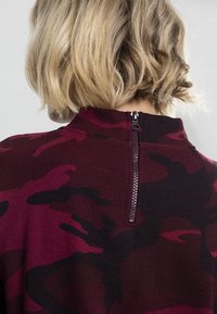 Sweter w moro w kolorze maroon i czarnym, z wysokim kołnierzem i widocznym zamkiem z tyłu. Miękka tkanina o teksturowanym wykończeniu.