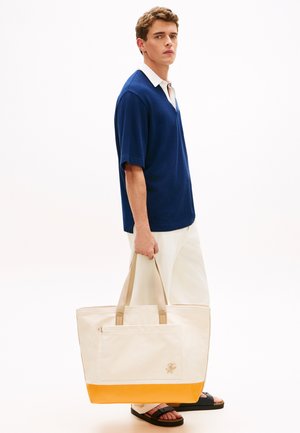 Ung mand iført en blå kortærmet polo og hvide bukser, der holder en stor beige taske med gul bund, iført sorte sandaler.