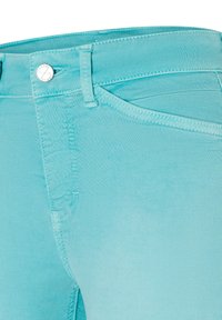 Gros plan d’un jean en denim turquoise montrant la ceinture, la boucle de ceinture, la poche avant et le bouton métallique avec texte gravé.