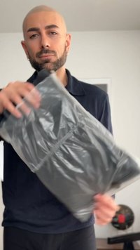 Vyras su barzda ir skutimosi galva laiko pilką plastiku apvyniotą dėžutę patalpose, ant paprasto fono.