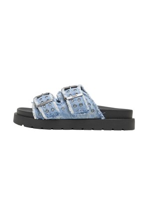 Sandalias tipo slide de mezclilla con tiras de doble hebilla, tela azul adornada con tachuelas de metal y una suela gruesa de goma negra.