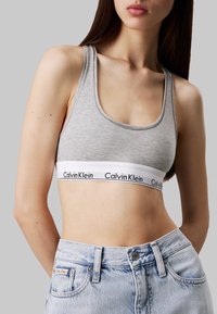 Sujetador deportivo de algodón gris con escote redondo y tirantes anchos, que presenta una banda blanca con "Calvin Klein" en letras negras. Combinado con jeans de un azul claro.