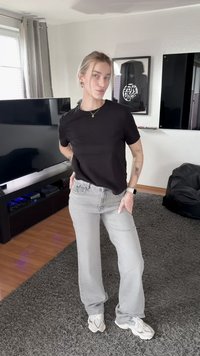 Jeune personne aux cheveux blonds, portant une chemise noire et un jean gris, se tient dans un salon avec télévision, œuvres d'art et fauteuil pouf.