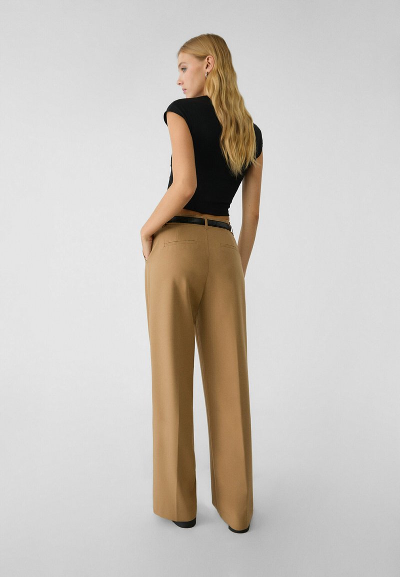 Donna con lunghi capelli biondi che indossa una maglietta nera a maniche corte infilata in pantaloni larghi color cammello a vita alta con una cintura nera.