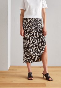 Jupe midi imprimée léopard en tissu beige et noir, dotée d'une fente sur le côté, associée à des mules noires à bout ouvert sur un parquet en bois.