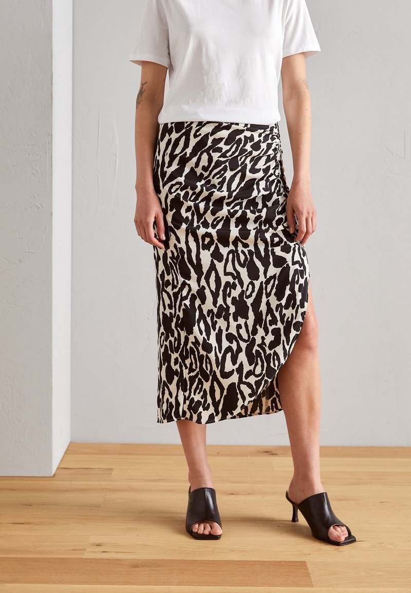 Jupe midi imprimée léopard en tissu beige et noir, dotée d'une fente sur le côté, associée à des mules noires à bout ouvert sur un parquet en bois.