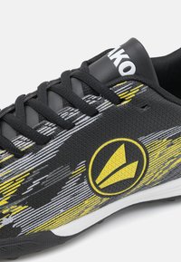 Zapatilla deportiva negra con diseño abstracto en amarillo y gris, que cuenta con una superficie texturizada, un sistema de cordones tradicional y un logo prominente en el lado.