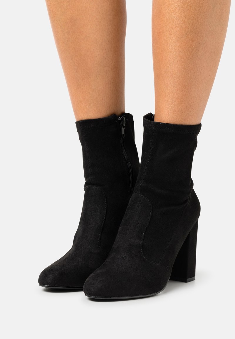 Bottes cheville en daim noir avec un talon bloc haut, dotées d'une fermeture éclair latérale et d'un shaft ajusté. Texture lisse avec des détails minimaux.