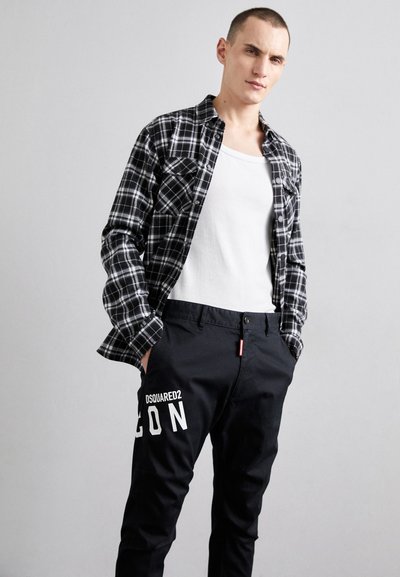 Black and white plaid flannel shirt, buttoned, over a white tank top. Black pants with a white logo and side pocket detail. Smooth fabric texture.