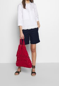 Vit bomullstopp med spetsdetaljer, marinblå shorts och en stor röd vävd väska. Svarta platta sandaler fulländar outfiten.