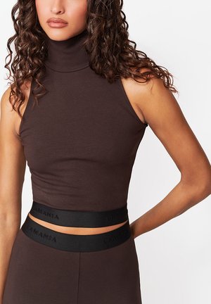 Kobieta ubrana w ciemnobrązowy bezrękawnik z golfem crop top oraz dopasowane, wysokie spodnie z czarnymi elastycznymi paskami z napisem "La Mania".