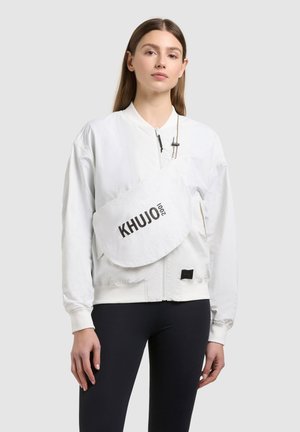 Woman wearing white zip-up jacket and white crossbody bag with "KHJO 2001" text, paired with black leggings against plain background.