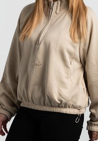 Beige džemperis ar stāvo apkakli, ar rāvējslēdzēju, elastīgu apakšmalu un sānu kabatu. Tam ir struktūrēta auduma virsma un diskrēta logo.