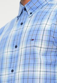 Μπλε πουκάμισο καρό με κολάρο που κλείνει με κουμπιά, σκούρα κουμπιά και μια μικρή λεπτομέρεια λογότυπου. Το σχέδιο περιλαμβάνει ανοιχτό μπλε και πράσινες γραμμές.