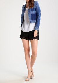 Jeansowa kurtka z kontrastową kieszenią, szara koszulka, czarne koronkowe szorty i beżowe sandały na wysokim obcasie. Czyste linie i wyraźne tekstury.