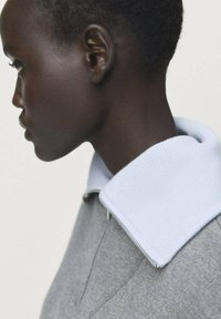 Perfil de una persona con el cabello corto que lleva una blusa gris con un gran cuello blanco doblado y un cierre visible.