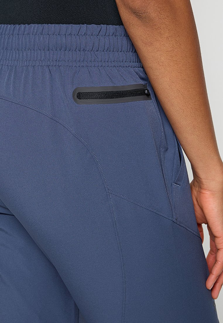 Personne portant un pantalon de sport bleu avec une taille élastique et une poche arrière zippée, se tenant debout avec la main droite détendue le long du corps.
