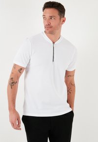 Bílá polo košile s krátkým rukávem a zipem u krku, vyrobená z texturované látky. Model má na sobě tmavé kalhoty a vystavuje tetování na paži.