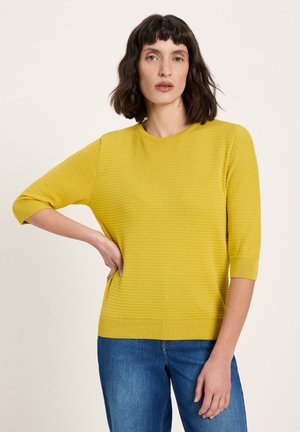 Woman with dark shoulder-length hair wearing a yellow ribbed elbow-length sleeve sweater and blue jeans, standing with one hand on hip.