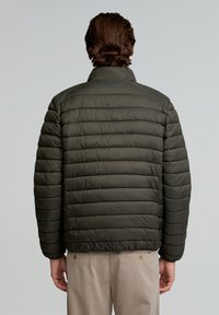 Chaqueta acolchada de un verde oscuro con diseño de acolchado horizontal, cuello alto y dobladillo elástico. Presenta un logo sutil cerca de la zona del cuello.