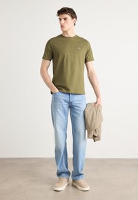 T-shirt à col ras du cou vert olive, jean bleu clair, chaussures beige, et tenant une veste marron clair. Tissu lisse avec un design ajusté.