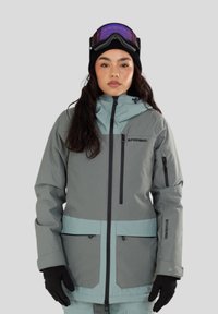 Grijze en teal ski-jas met een ritssluiting aan de voorkant, zwarte hardware en pocketdetails. Gecombineerd met een zwarte gebreide muts en paarse skibril.