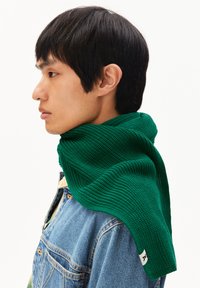 Écharpe verte texturée drapée sur une veste en denim, mettant en valeur un motif en nid d'abeille et une étiquette blanche. L'écharpe est bien mise en avant contre un fond clair.