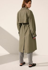 Trench coat verde oliva con vita cinta, dettaglio con pattina sul retro e bottoni sui polsini. Indossato sopra pantaloni grigi a gamba larga e sandali neri.